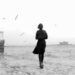 A woman walks along a waterfront in black and white, with birds flying around her.